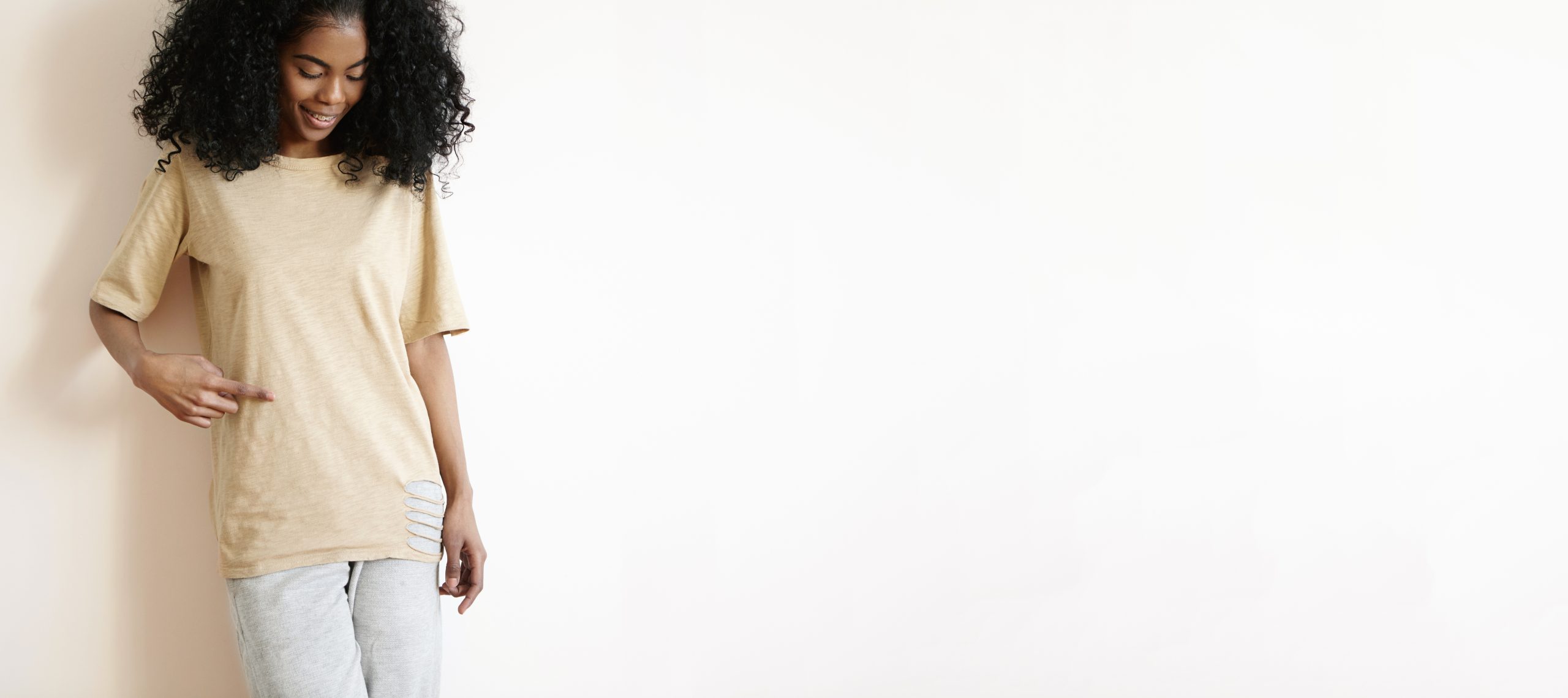 Studio portrait of attractive young African female model with stylish curly haircut dressed casually smiling cheerfully, showing copy space for your print on her t-shirt with finger. Horizontal